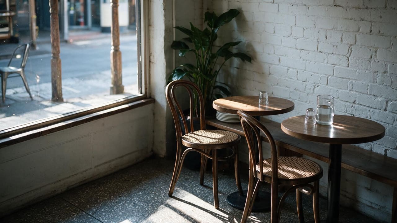 An empty cafe corner before opening, with two bentwood chairs, small round tables and morning light across a terrazzo floor.
